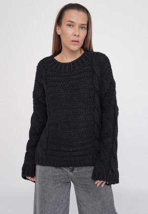 Black knitted sweater featuring a textured cable pattern, round neckline, and long, wide sleeves. Paired with light gray denim pants.