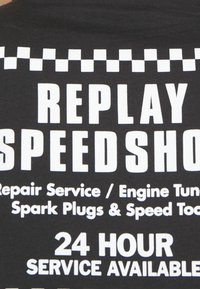 Černá látka s tučným bílým textem. "REPLAY SPEEDSHOP" nahoře, s dalšími podrobnostmi o službách níže. Klobouček s šachovnicovým vzorem nad textem.