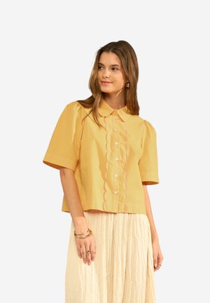 Kvinde iført en gul bluse med korte ærmer med bølgede knapper foran og et beige plisseret skørt, tilbehør med guldsmykker.