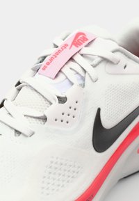 Baskets blanches avec un dessus texturé, swoosh Nike noir, accent rose sur l'étiquette des lacets, et un design principalement en mesh pour la respirabilité.