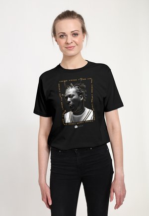 Kvinde iført sort t-shirt med et sort-hvidt portræt af en mand med flettet hår og gyldne accenter omkring billedet.