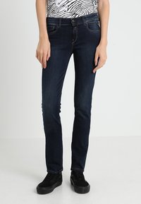 Jeans skinny blu scuro in denim, caratterizzati da un classico design a cinque tasche e una texture liscia, abbinati a sneakers nere.