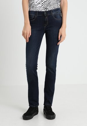 Jean skinny bleu foncé en denim, présentant un design classique à cinq poches et une texture lisse, associés à des baskets noires.