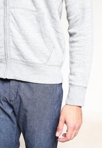 Hombre con una sudadera con cremallera gris claro y pantalones azul oscuro, con la mano relajada a un lado contra un fondo liso.