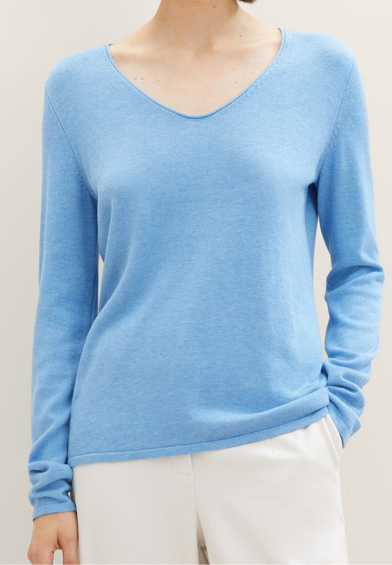 Frau trägt einen hellblauen, langärmeligen V-Ausschnitt-Pullover und weiße Hose, eine Hand in der Tasche, beige Hintergrund.