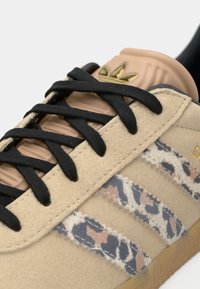 Beige Wildledersneaker mit schwarzen Schnürsenkeln, die mit Leopardenmuster-Streifen und einer hellbraunen Gummisohle ausgestattet sind. Goldene Markenlogo auf der Zunge.