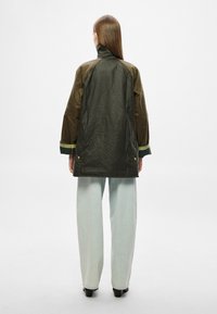 Πράσινο και καφέ αδιάβροχο μπουφάν με ψηλό γιακά, μακριά μανίκια και αντίθετα μανσέτα. Συνδυάζεται με ανοιχτό μπλε τζιν παντελόνι φαρδιάς γραμμής.