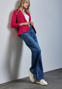 Veste cintrée rose sur un débardeur blanc, associée à un jean évasé bleu et des baskets blanches. Textures lisses, design décontracté et couleurs vives.