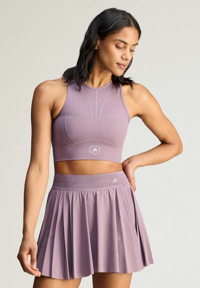 Woman wearing a lilac sports crop top and pleated skirt, standing with one hand on hip and looking down to the side.