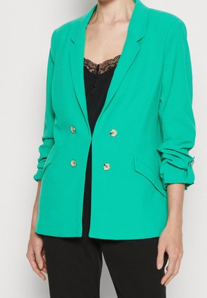 Femme portant un blazer croisé bleu sarcelle aux manches retroussées, par-dessus un haut noir orné de dentelle, et un pantalon noir.