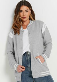 Grijze jersey bomberjack met witte gestreepte accenten op de mouwen, drukknoopsluiting en zijzakken. Ggedragen over een wit t-shirt en jeans.