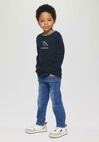 Marineblaues Langarmshirt mit grünen Akzenten, grafischem Text und blauen Jeans. Weiße Turnschuhe mit grauen und roten Details.