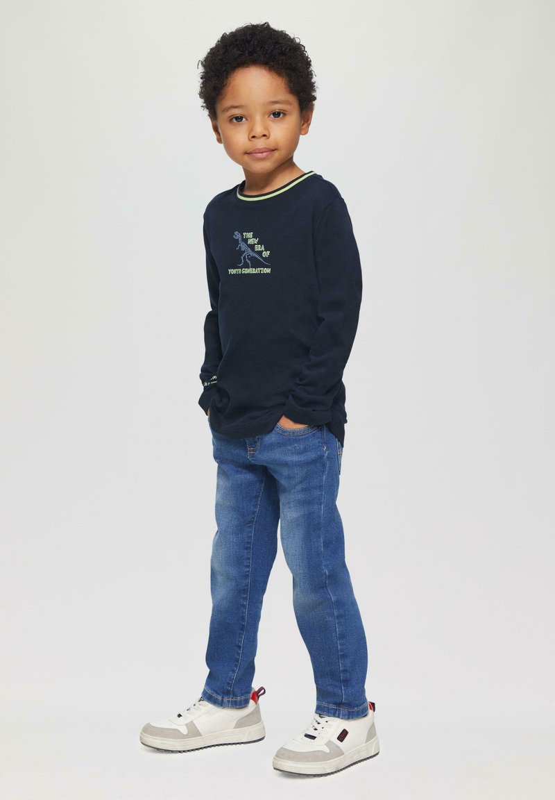 Marineblaues Langarmshirt mit grünen Akzenten, grafischem Text und blauen Jeans. Weiße Turnschuhe mit grauen und roten Details.