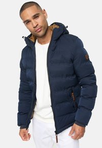 Man som bär en marinblå vadderad jacka med huva över en vit skjorta och vita byxor, stående mot en enfärgad bakgrund.