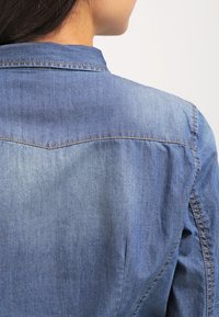 Jeansskjorta i en enhetlig ljusblå färg, med knäppningkrage och detaljerad sömnad längs axlarna och bakstycket.