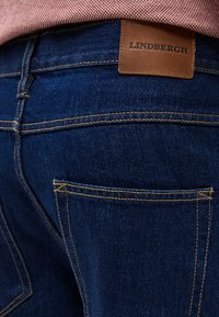 Πίσω τσέπη σκούρου μπλε τζιν με καφέ δερμάτινη ετικέτα μάρκας Lindbergh και ορατή ραφή, φορεμένα με ροζ υφασμάτινο τοπ.