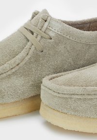 Scarpe in suede di colore beige chiaro con una superficie texturizzata. Presentano un design semplice con punta rotonda e suola in gomma marrone. I lacci accentuano la parte superiore.