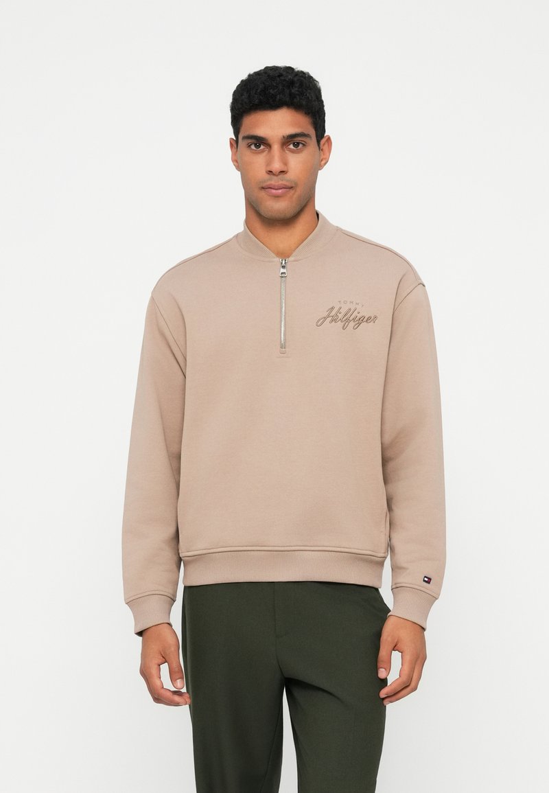 Béžová mikina se zipovým límcem, na kterém je vyšívaný nápis "Tommy Hilfiger" v kontrastní barvě. Měkká tkanina a žebrované manžety.