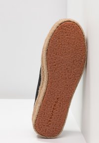 Semelle de chaussure espadrille noire avec une base en caoutchouc brun texturé et des détails en corde de jute le long du bord, présentée sur un fond blanc.