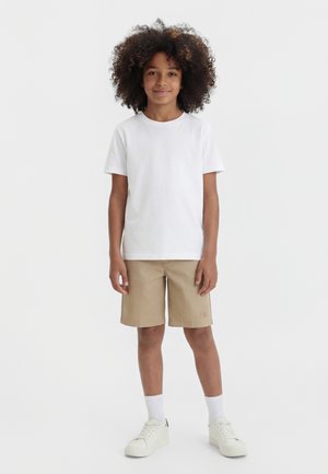 Mladá osoba kučeravými vlasmi, oblečená v bielom tričku, béžových šortkách, bielych ponožkách a bielych teniskách, stojaca pred jednoduchým pozadím.