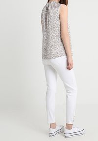 Blouse sans manches à motif floral beige et gris, associée à un pantalon blanc slim et des baskets blanches avec des accents noirs.
