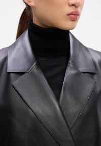 Musta nahkblazer laiade kraega, sile tekstuur ja kohandatud lõige, kantud musta roll-neck'iga. Puhtne ja minimalistlik disain.