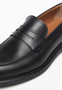 Mocassino in pelle nera con finitura liscia, caratterizzato da una banda cucita sulla tomaia e una punta arrotondata. La suola è piatta e leggermente strutturata.