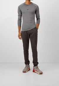 Person wearing gray long-sleeve Nike top, dark jogger pants, and gray and red Nike sneakers standing with one hand in pocket.