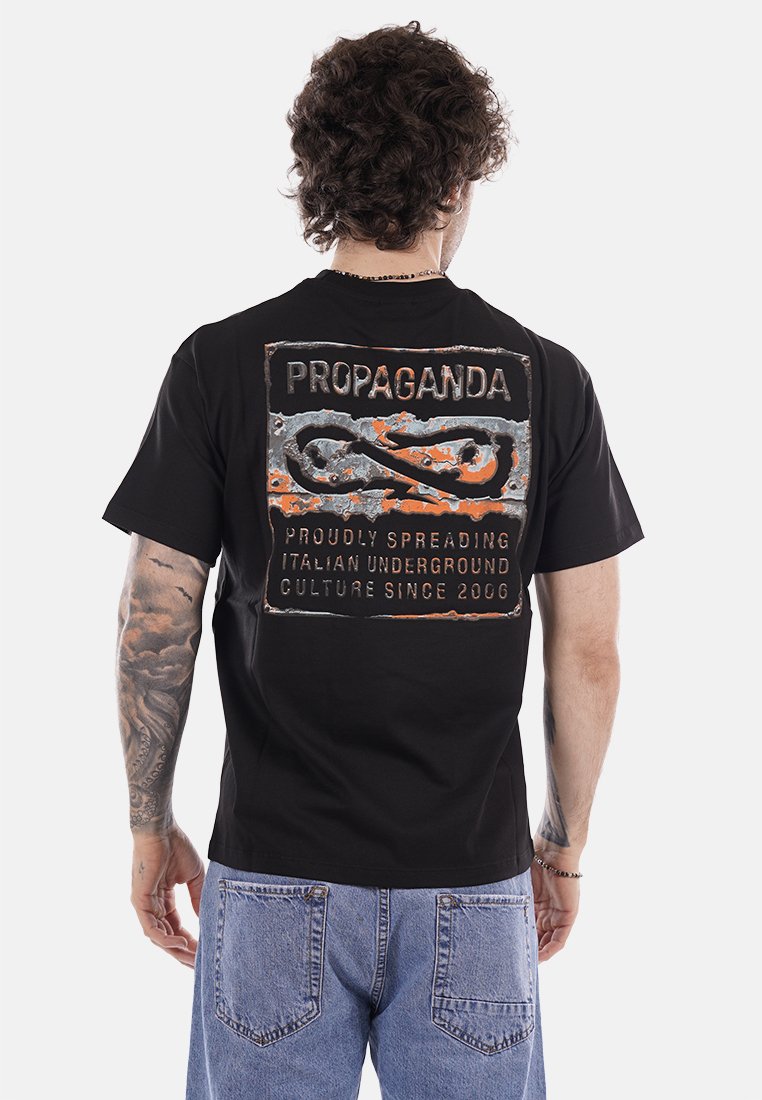 Uomo con capelli ricci che indossa una maglietta nera con la scritta "PROPAGANDA" e testo grafico arancione-grigio sul retro, abbinata a jeans blu.