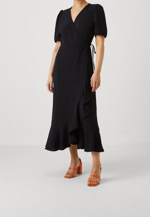 Kobieta w czarnej sukience kopertowej z bufkami i marszczonym dołem, ubraną w pomarańczowe sandały na obcasie, stojąca na jasnym, jednolitym tle.