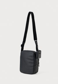 Svart rektangulær crossoverveske med justerbar stropp og liten festet glidelåslomme hengende mot hvit bakgrunn.