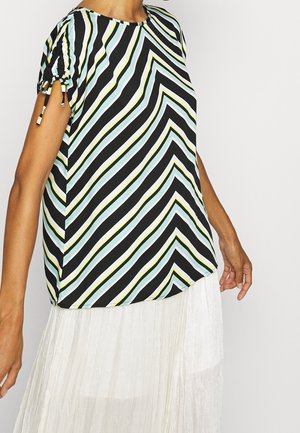 Vrouw draagt een top met korte mouwen met diagonale zwarte, witte, gele en lichtblauwe strepen, gecombineerd met een witte geplooide rok.