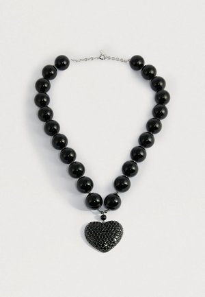 Melna krellīte ar lielām spīdīgām pērlēm un teksturētu melnu sirds formas kulonu uz sudraba ķēdītes aizdares.