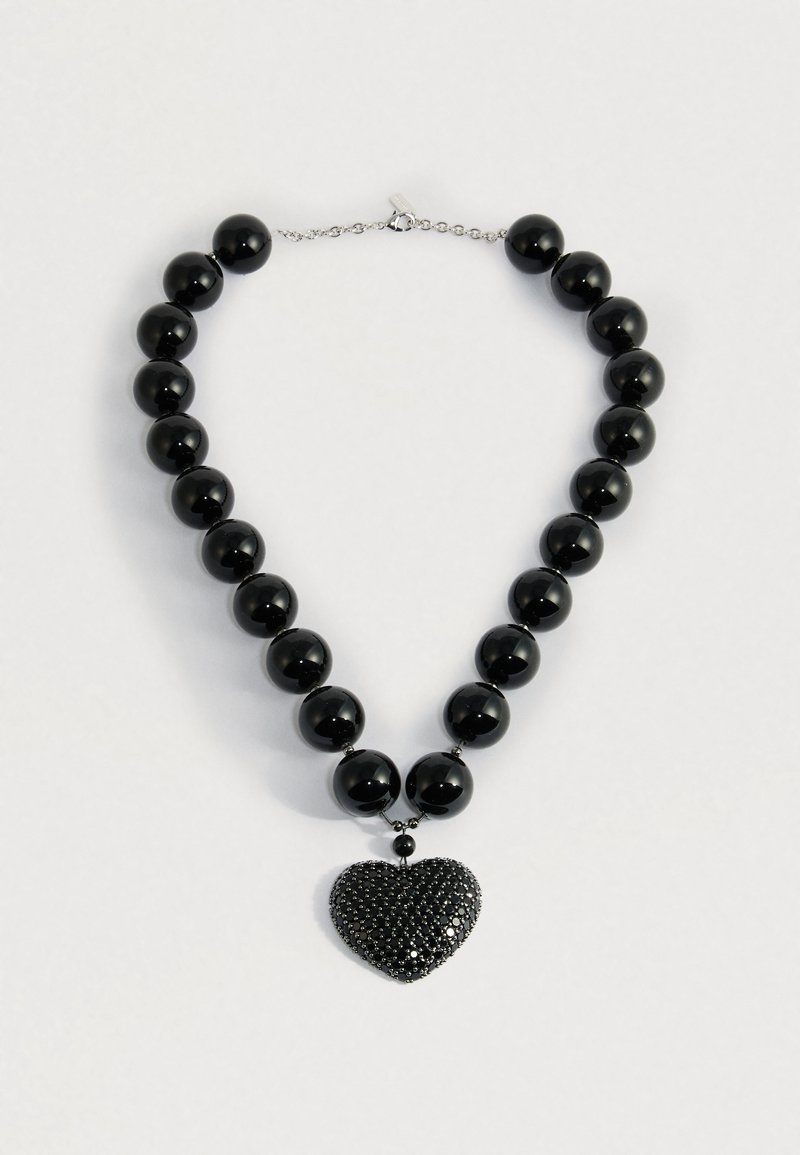 Melna krellīte ar lielām spīdīgām pērlēm un teksturētu melnu sirds formas kulonu uz sudraba ķēdītes aizdares.