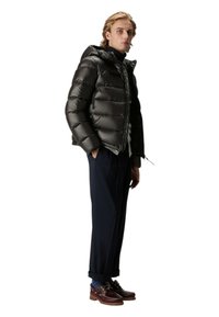 Zwarte gewatteerde jas met capuchon, glanzende afwerking, ritssluiting en elastische manchetten. Draagt over een coltrui en gecombineerd met marineblauwe broek en bruine schoenen.