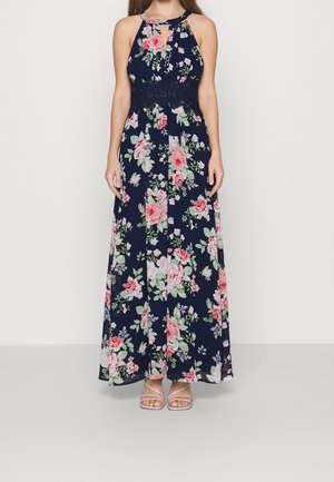 Kvinde iført en ærmeløs, navyblå maxikjole med rosa og hvide blomsterprint og blonder i taljen, kombineret med nøgenfarvede sandaler med stropper.