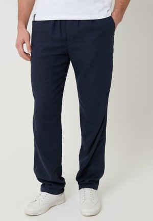 Uomo che indossa pantaloni casual blu navy e sneakers bianche, in piedi con una mano in tasca davanti a uno sfondo semplice.