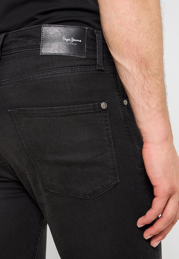Jeans de mezclilla negra con un parche de cuero que presenta el logo de la marca. Los bolsillos traseros tienen detalles de costura y remaches de metal. Tejido texturizado.