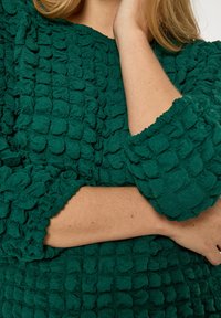 Zelený textúrovaný top s plyšovým povrchom a jedinečným vystúpeným vzorom. Rukávy sú na manžetách nazbierané pre väčší detail.