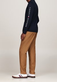 Langärmeliges marineblaues Hemd mit „Tommy Hilfiger“-Logo in Weiß auf dem Ärmel, kombiniert mit sandfarbenen Chinos und weißen Turnschuhen mit bunten Streifen.