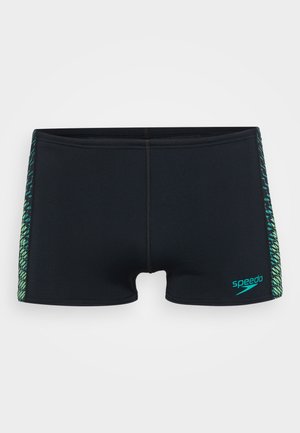 Melnas peldbikses ar ciešu piegriezumu, kurām sānos ir zaļi un zili ģeometriski raksti un neliels "Speedo" logotips priekšā.
