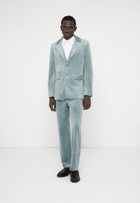 Svileni komplet u pastelno tirkiznoj boji. Jednoredni sako s reverima s urezom, dva prednja džepa, uparen s odgovarajućim hlačama i crnim cipelama.