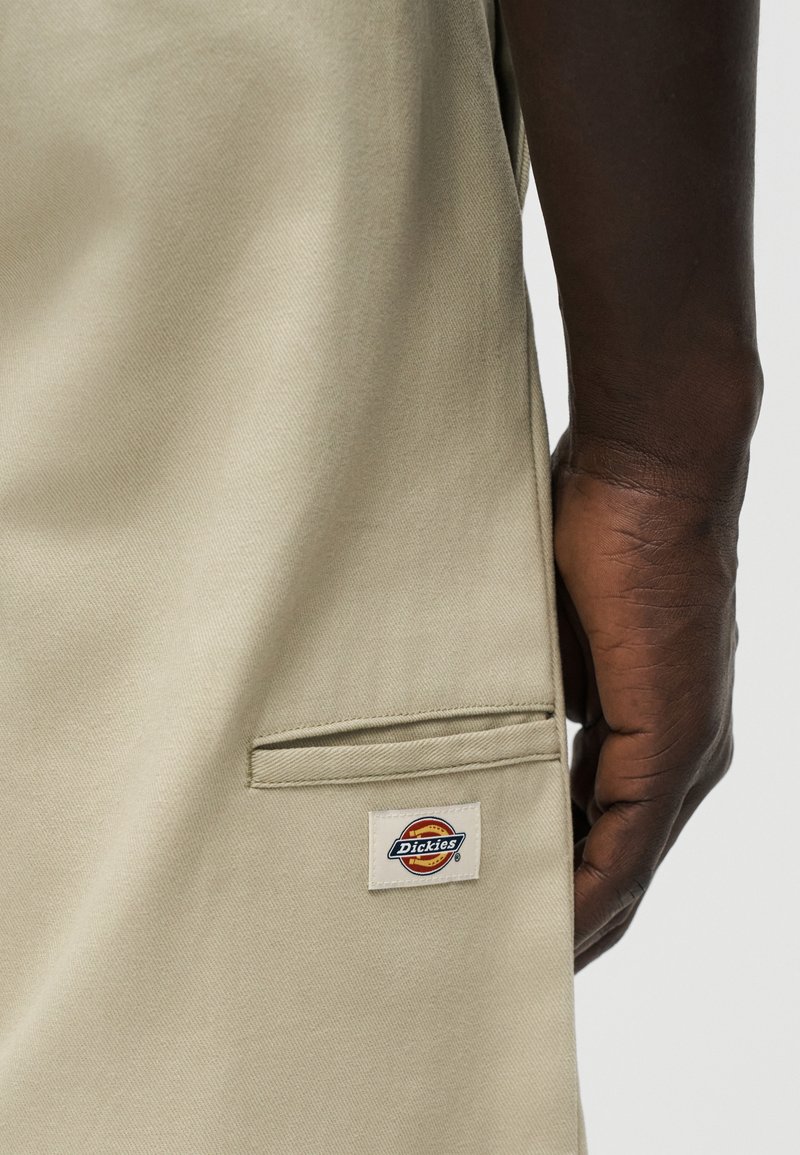 Pantalon Dickies beige avec détail de poche passepoilée sur le côté près de la hanche, une main à peau foncée reposant à côté.