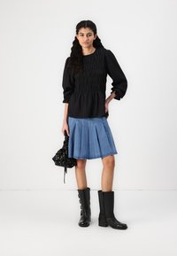 Black smocked blouse, light blue pleated denim skirt, and black knee-high boots. Model holds a black floral bag, with a plain white background.