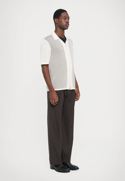 Short-sleeve knit shirt in light beige with a textured pattern, paired with dark brown trousers and black shoes. Button-down front and collar.