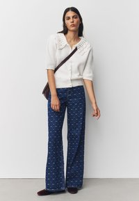 Biały dziany kardigan z falistym kołnierzem, w połączeniu z niebieskimi rozszerzanymi dżinsami z wzorzystym motywem. Modelka trzyma brązową torebkę.