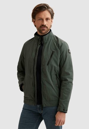 Mann mit kurzen braunen Haaren und Bart, trägt eine dunkelgrüne Reißverschlussjacke und blaue Jeans, steht mit einer Hand in der Tasche.