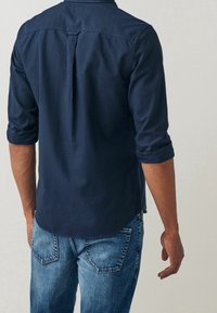 Chemise bleu marine avec un pli au dos, manches longues retroussées et ourlet arrondi. Portée avec un jean en denim bleu clair avec des poches arrière.