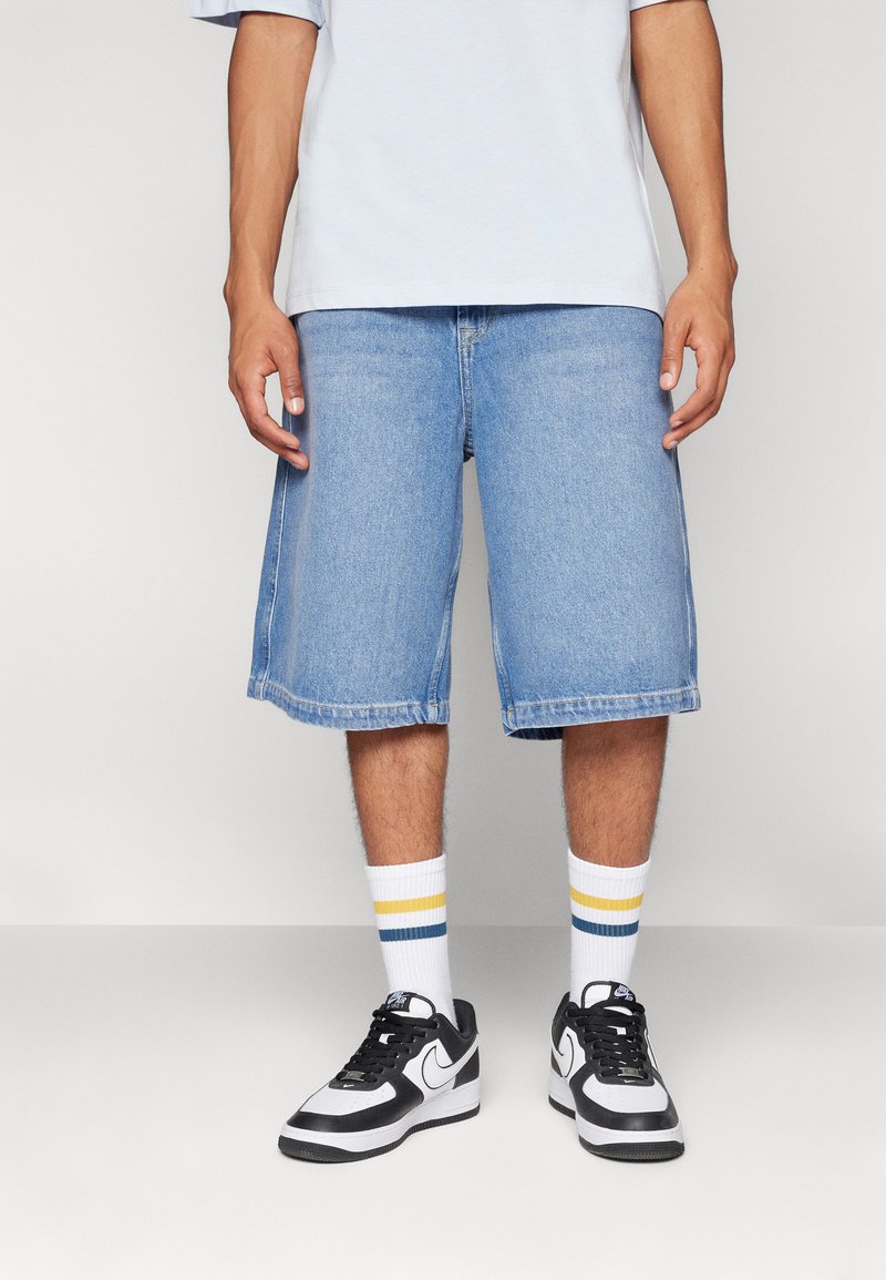 Lyseblå denimshorts med en afslappet pasform, der har en frynset kant, båret med hvide sokker med blå og gule striber og sorte sneakers.