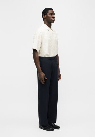 Άνδρας που στέκεται φορώντας ένα κοντομάνικο λευκό πουκάμισο με κουμπιά, σκούρο, φαρδύ παντελόνι και μαύρα παπούτσια σε ένα απλό φόντο.